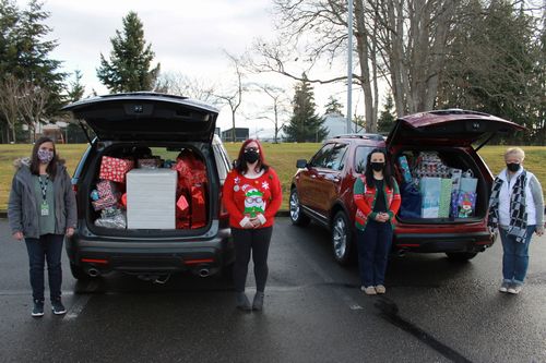 Bethel School District Communication coordinators and holiday-adorned CMA employees stand in front of the two cars full of gifts